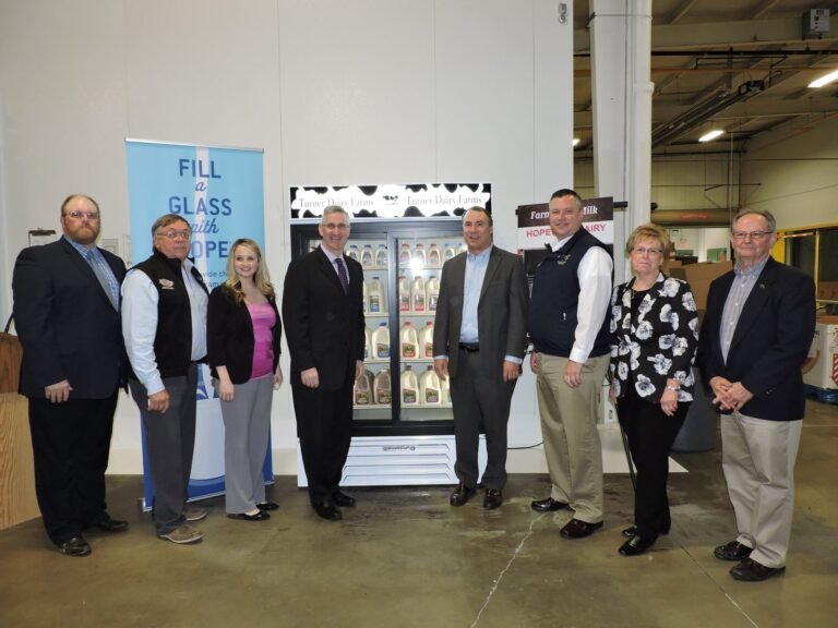 Eight people stand indoors in front of a refrigerated display of milk bottles. A blue sign says “Fill a Glass with Hope.” The group is dressed in business or business-casual attire, posing for a photo.