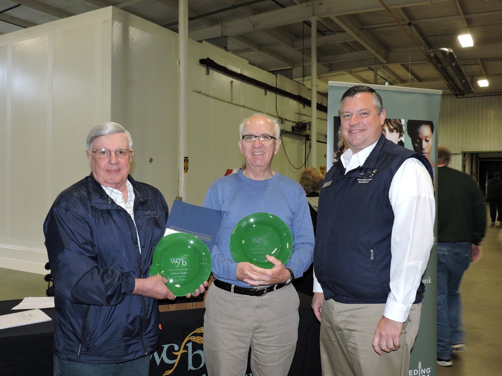 Three men stand indoors; two hold green plates with wyfb printed on them, smiling at the camera. The third man stands to the right, wearing a navy vest. Industrial setting in the background.