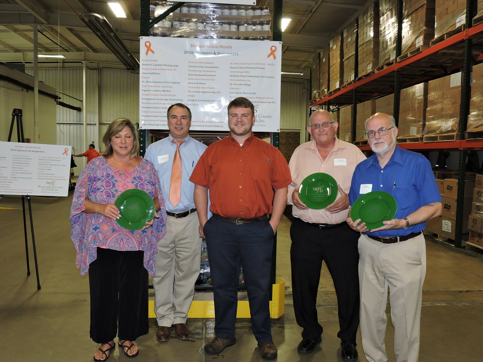 Five people stand indoors in a warehouse, posing for a group photo. Three men hold green hard hats. Boxes are stacked on shelves behind them, and informational posters are visible in the background.
