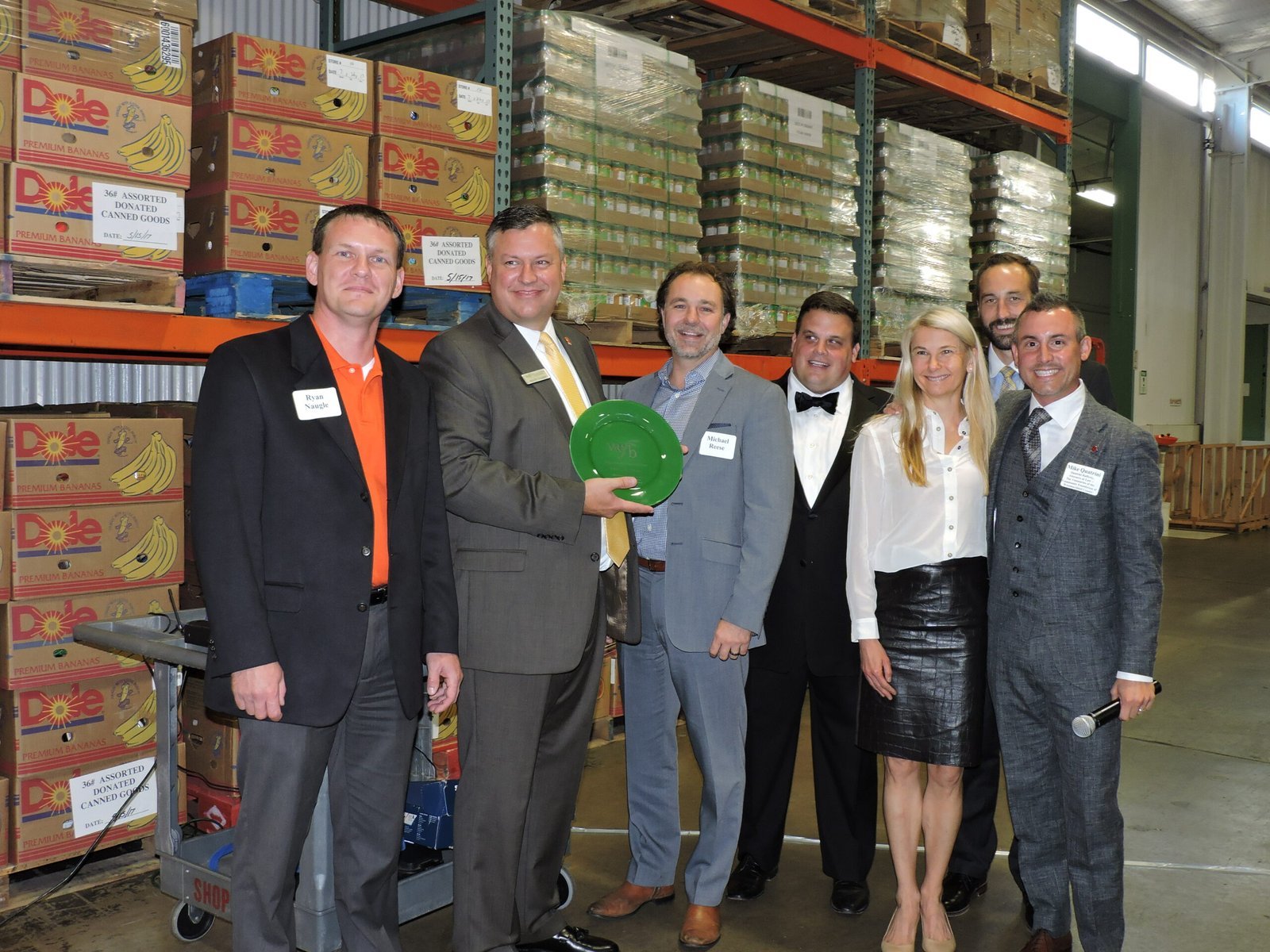 Seven people in business attire stand together smiling in a warehouse with shelves of stacked boxes. One person holds a green plate award. Name tags are visible on their jackets. Pallets and carts are seen in the background.