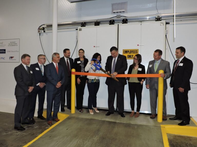 A group of people in business attire stand in front of large white doors as a man and woman cut an orange ribbon with oversized scissors at a ribbon-cutting ceremony. A sign on the door reads Employees Only.