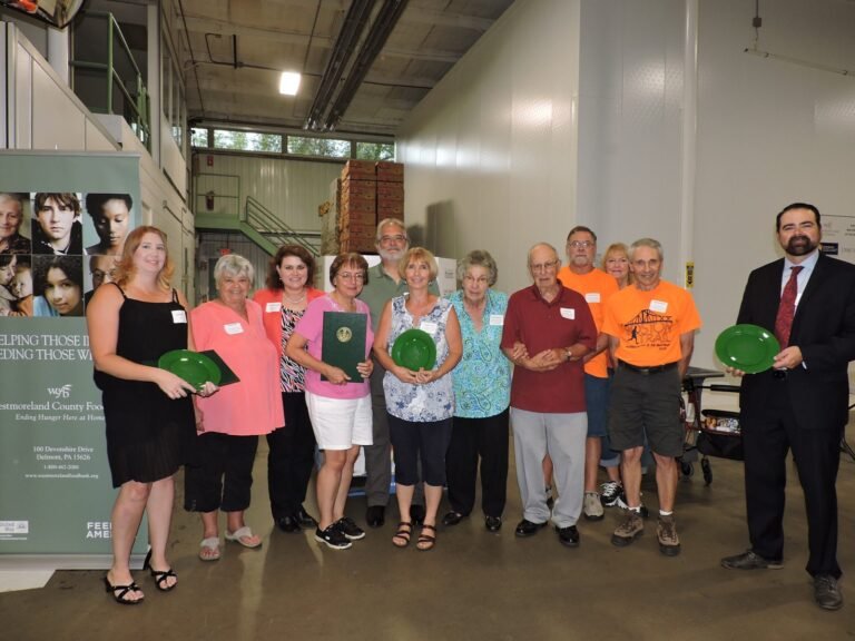 A group of people stand indoors, some holding green plates, posing for a photo. They are smiling, and a banner about a food bank is visible in the background along with storage shelves.