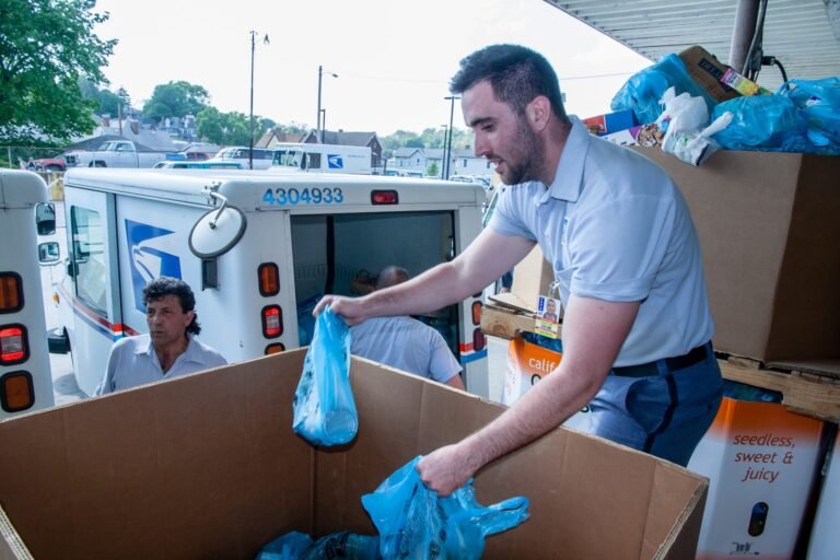 A man in a postal uniform places blue plastic bags filled with donations into a large box, while other postal workers and delivery trucks are visible in the background.
