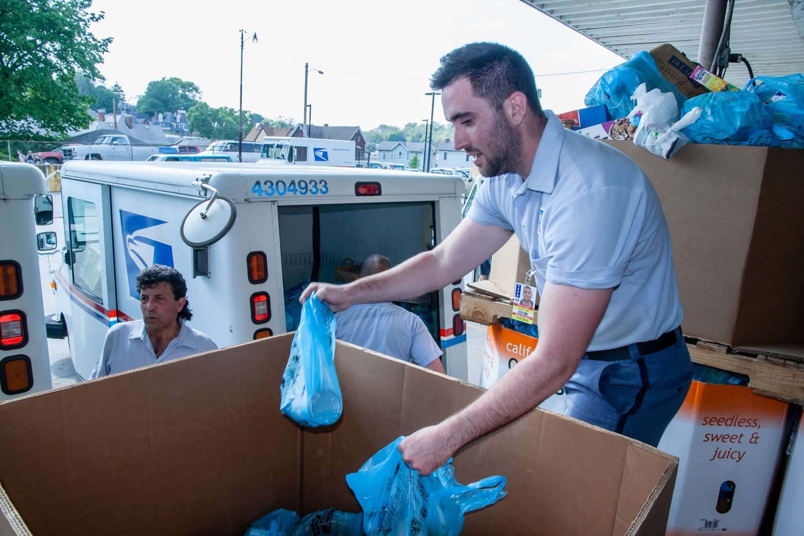 A man in a postal uniform places blue plastic bags filled with donations into a large box, while other postal workers and delivery trucks are visible in the background.