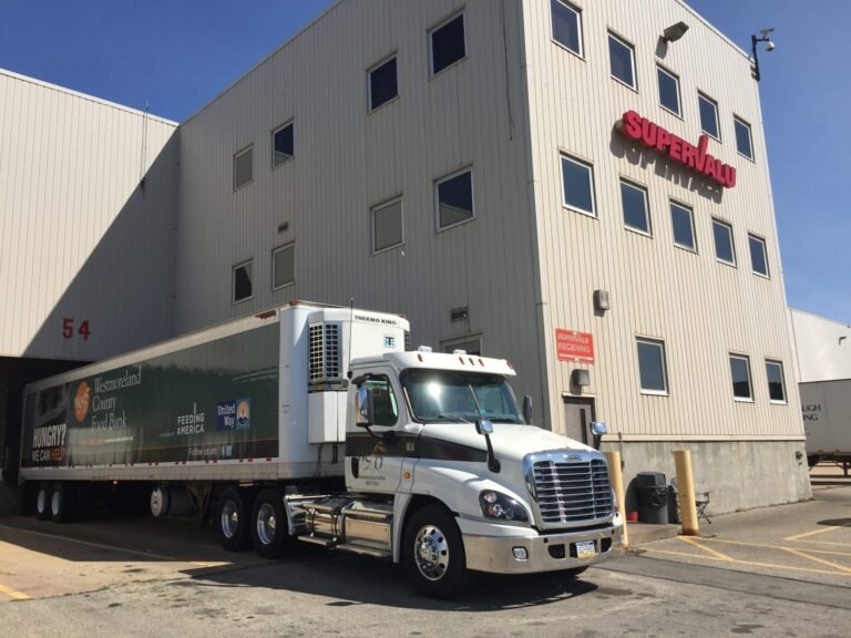 A white semi-truck with a refrigerated trailer is parked at a loading dock outside a large beige SuperValu warehouse building under a clear blue sky.