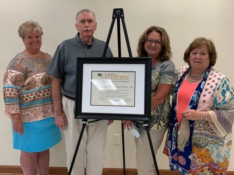 Four adults stand together indoors, smiling, with a framed certificate of excellence displayed on an easel between them. They are casually dressed and posed against a plain wall.