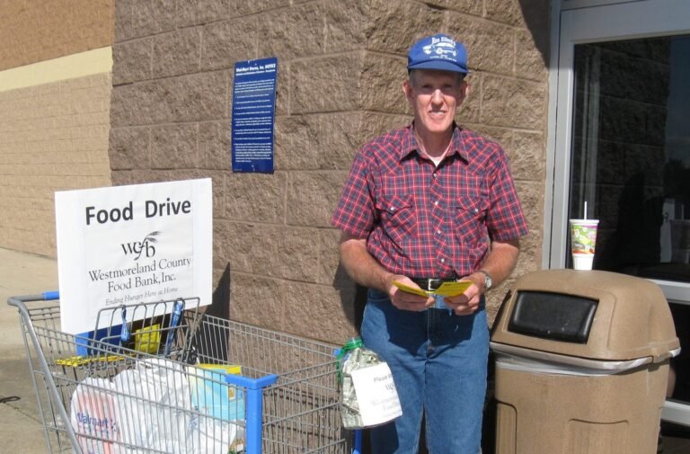 A man in a blue cap and plaid shirt stands by a shopping cart labeled Food Drive for Westmoreland County Food Bank outside a store, holding flyers next to a trash can and a drink.