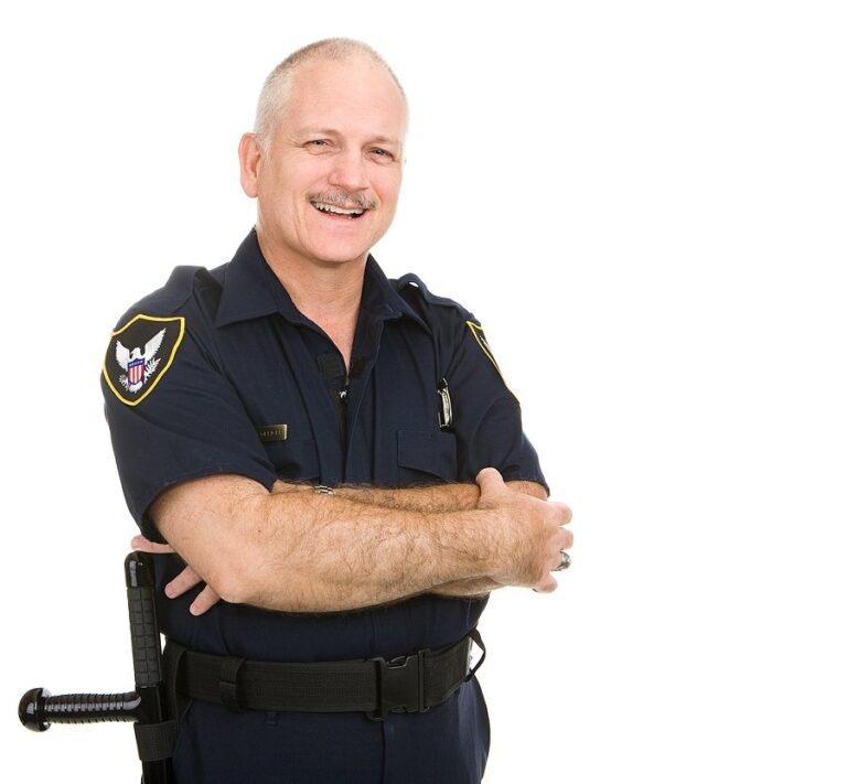 A smiling police officer in a navy blue uniform stands with arms crossed against a plain white background. The officer has short hair, a mustache, and wears a utility belt with equipment.