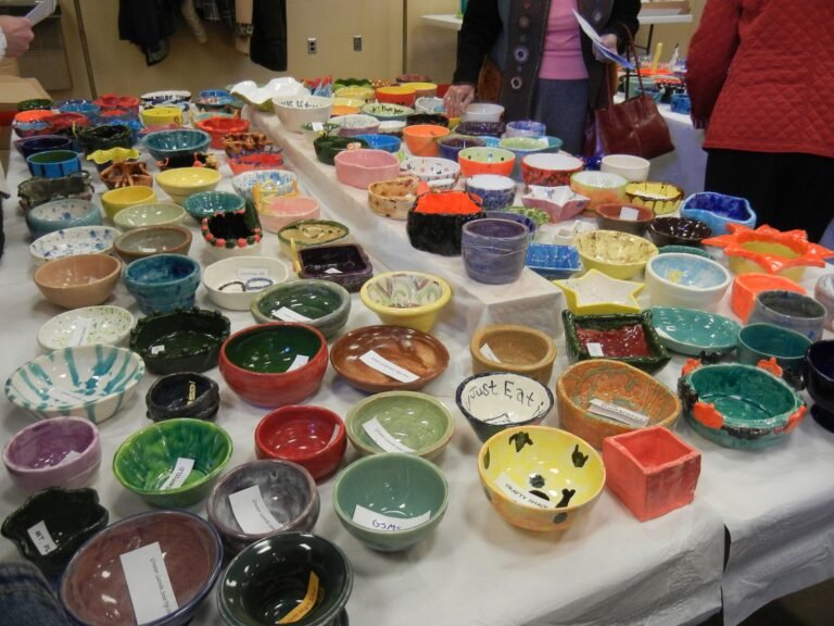 A large display of colorful, handmade ceramic bowls, plates, and dishes are arranged on tables covered with white cloths at an indoor art or craft fair. People are browsing the items in the background.