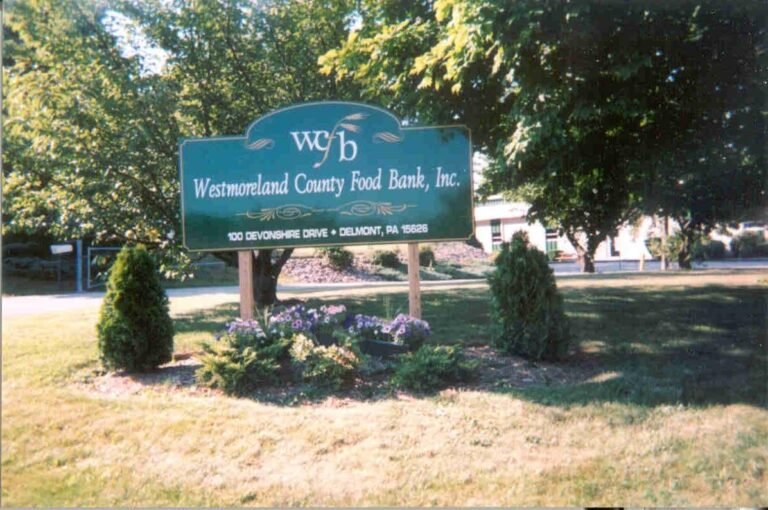A green sign reads Westmoreland County Food Bank, Inc., 100 Devonshire Drive, Delmont, PA 15626, standing on a grassy area with bushes and trees in the background.