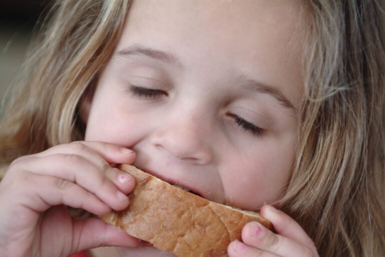A young child with long, light brown hair and closed eyes takes a big bite of a sandwich or piece of bread, holding it with both hands.