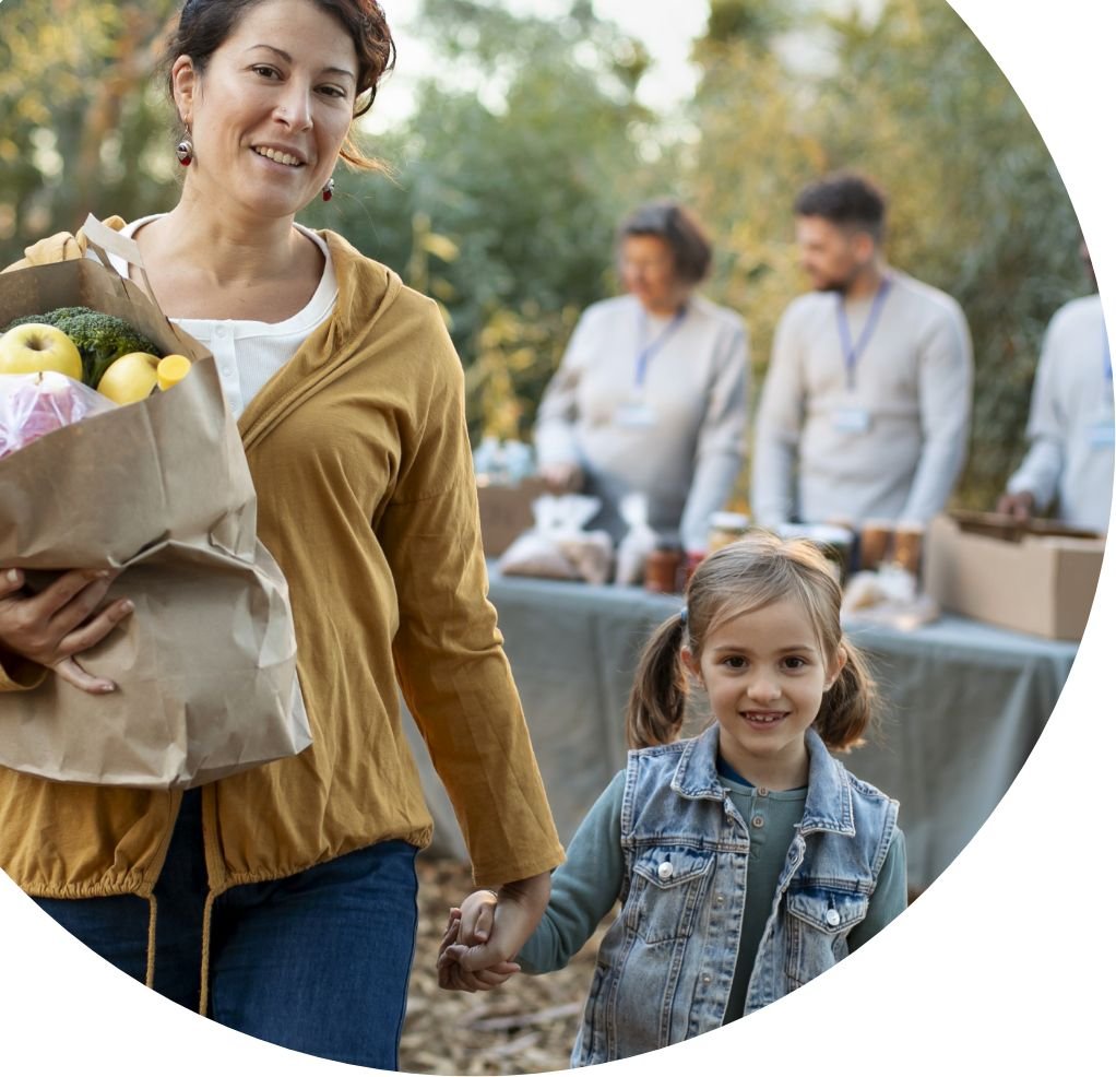 A woman with a grocery bag and a young girl holding her hand walk outdoors. In the background, people gather at a table with food items, suggesting a Feeding America food bank or pantry distribution event in the community.