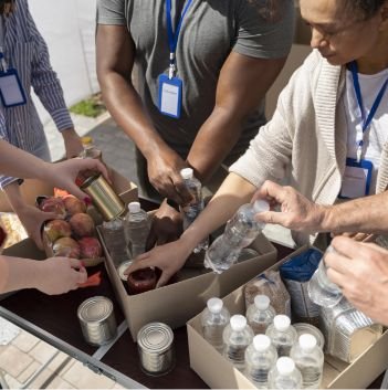 Four people wearing name tags pack bottled water, canned goods, apples, and packaged food into boxes on an outdoor table, likely volunteering for a food bank or pantry to provide food assistance to those in need.