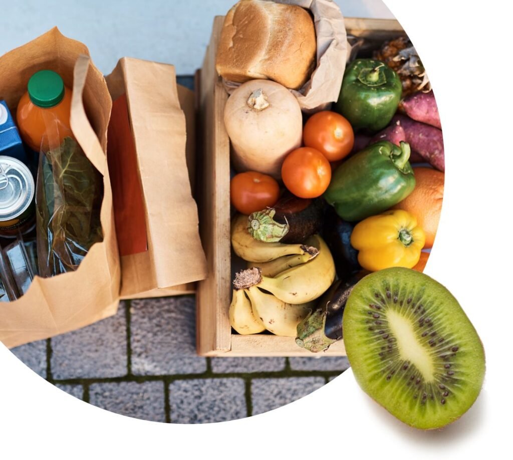 A wooden crate filled with fresh fruits and vegetables sits next to a grocery bag containing bottles and cans, resembling a food bank pantry; a sliced kiwi is placed in the foreground.