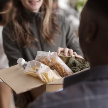 A person hands a box of food items, including pasta, bread, and a bottle, to another person at a food bank. Both individuals are smiling and interacting, suggesting a food donation or charitable exchange.