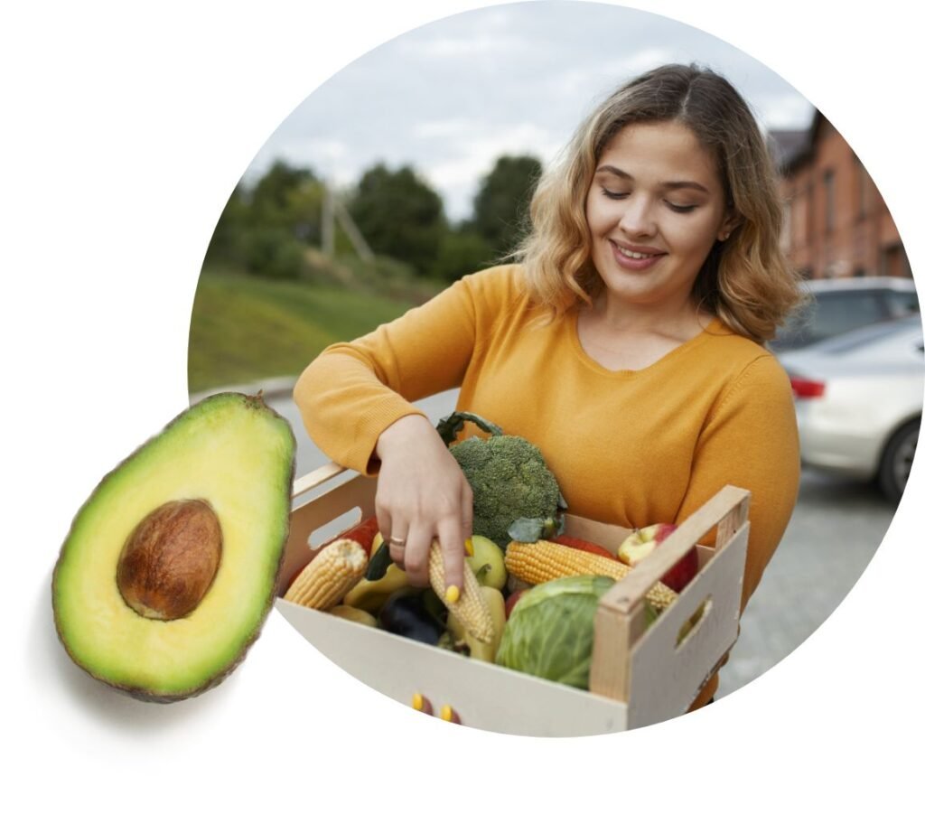 A woman in a mustard yellow top smiles while holding a wooden crate filled with various vegetables, showcasing the fresh produce often provided by a food bank. An avocado, cut in half, is displayed in the foreground beside the circular photo.