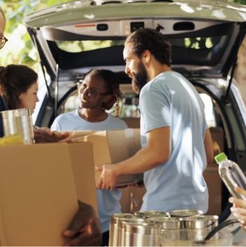 Volunteers in light blue shirts unload boxes and canned goods from a car, working together outdoors to provide food help during a community food drive for the local pantry and Feeding America.