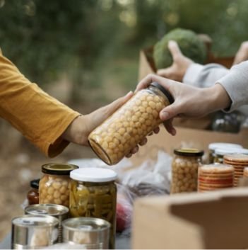 A person hands a glass jar filled with chickpeas to another at an outdoor food bank distribution table lined with boxes and jars of preserved food, offering much-needed food help to the community.