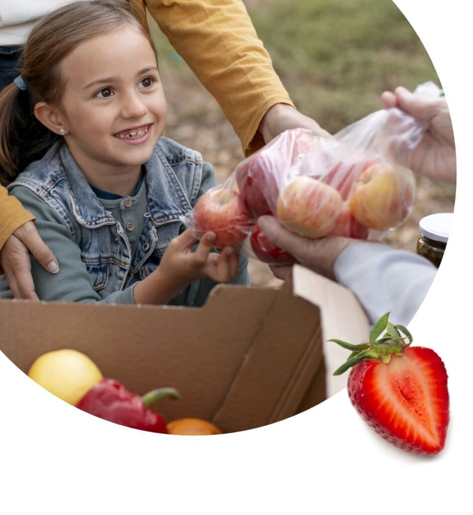 A smiling young girl receives a bag of apples from an adult at a food bank, with a cardboard box of assorted fruit nearby and a strawberry half in the corner.