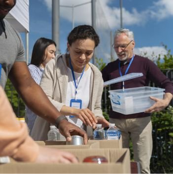A group of volunteers sorts food and water bottles into boxes outdoors on a sunny day, providing food help. One person holds a clear plastic bin while others organize supplies on a table for the local pantry.