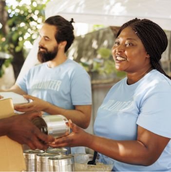 Two volunteers in blue shirts are smiling and handing out canned food at a food bank outdoors. One woman is passing a can to someone, while a man stands beside her, holding another can. Trees and a tent are visible in the background.
