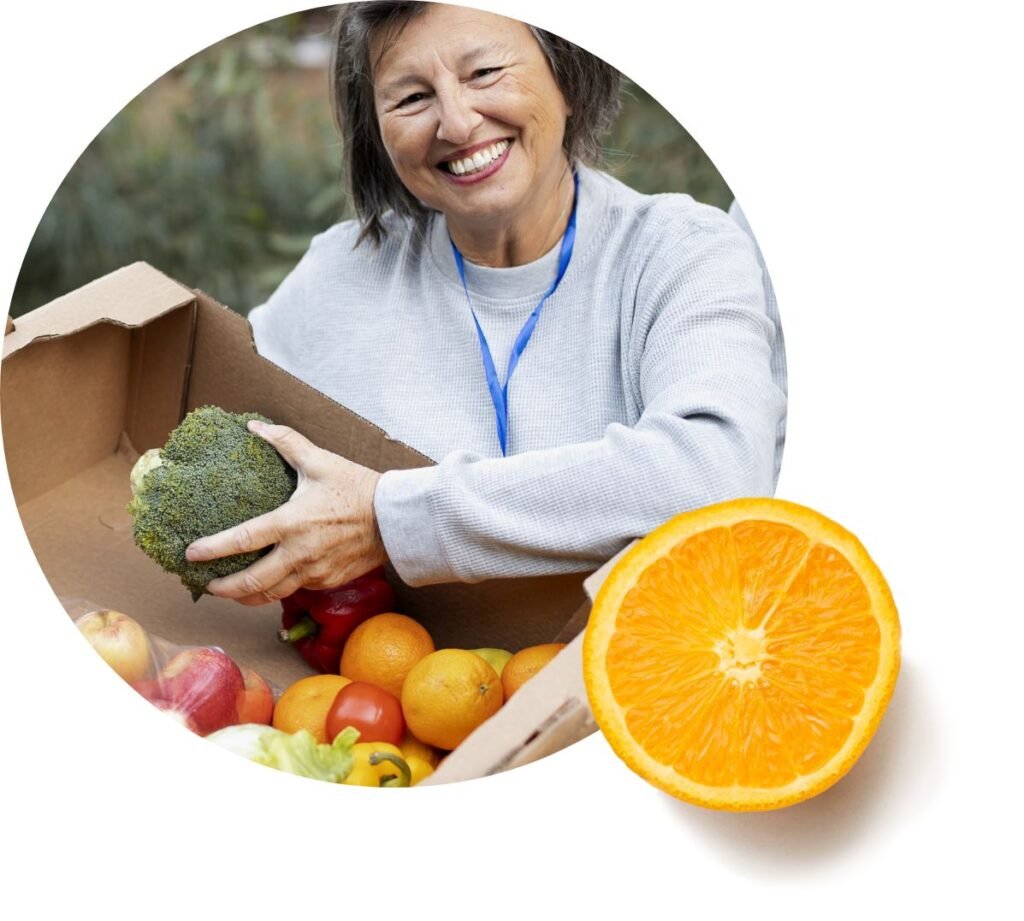 Smiling woman holding a box of fresh fruits and vegetables, including broccoli, apples, oranges, and tomatoes. A halved orange is placed in the foreground, symbolizing food assistance from Feeding America.