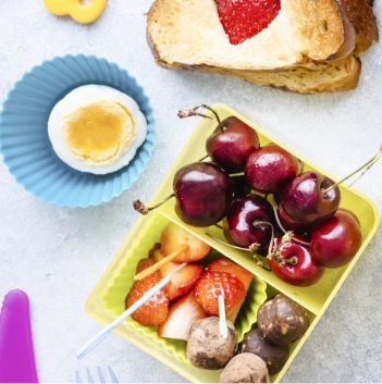 A lunchbox with fresh cherries, cut strawberries, and chocolate-covered treats sits beside a peeled hard-boiled egg in a blue cup and toast topped with sliced strawberry—perfect for sharing ideas about food assistance or Feeding America on a light surface.