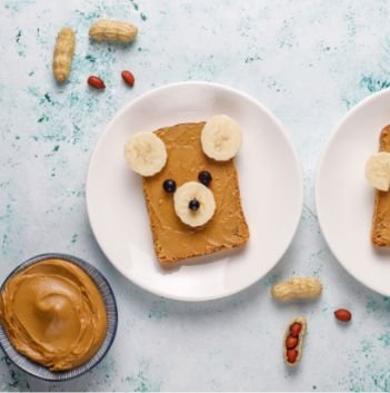 A slice of bread with peanut butter, decorated with banana slices and blueberries to look like a bear face, sits on a white plate—reminding us how food help from organizations like Feeding America can brighten every meal. Peanuts and a bowl of peanut butter are nearby.