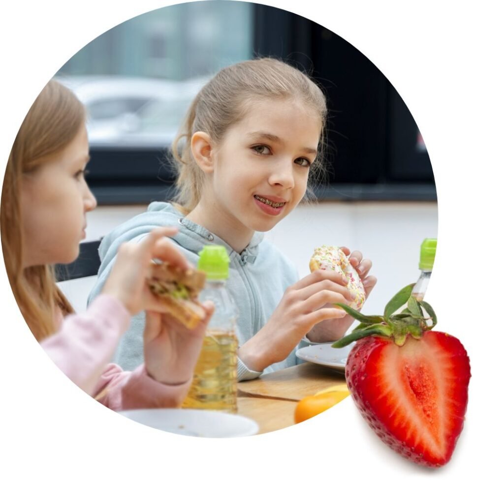Two girls sit at a table enjoying snacks and drinks, with one girl smiling at the camera while holding a sprinkled cookie. A large strawberry sits in the foreground, highlighting the joy food assistance can bring to families.