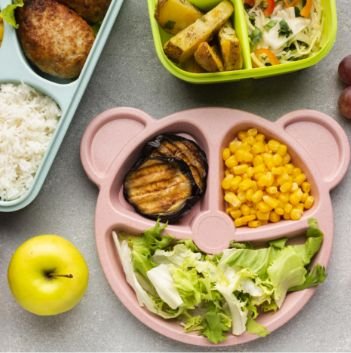 A pink bear-shaped plate with grilled eggplant, corn, and lettuce sits next to a yellow apple, rice, grapes, and another container with roasted potatoes and salad—perfect for anyone seeking food help or pantry inspiration.