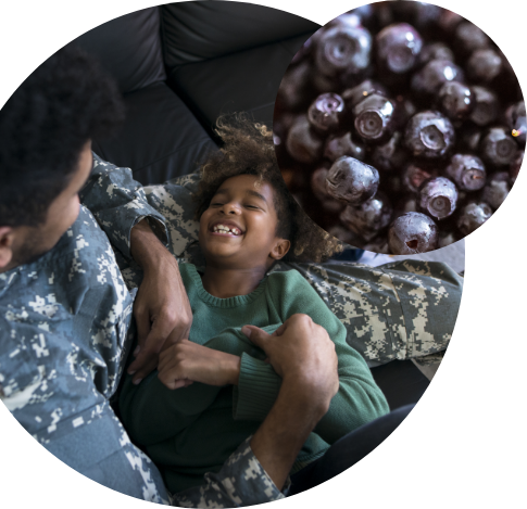 A person in military uniform playfully tickles a smiling child on a couch; a circular inset shows fresh blueberries, highlighting the nutritious impact of Feeding America’s food assistance programs.