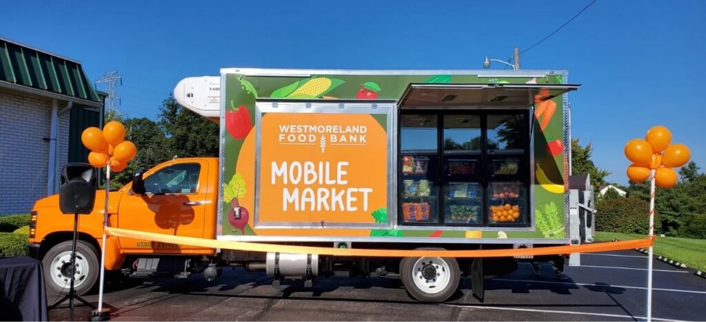 A brightly colored food truck labeled Westmoreland Food Bank Mobile Market offers fresh produce and food assistance outdoors. Orange balloons and ribbon decorate the area, suggesting a pantry celebration or special event.